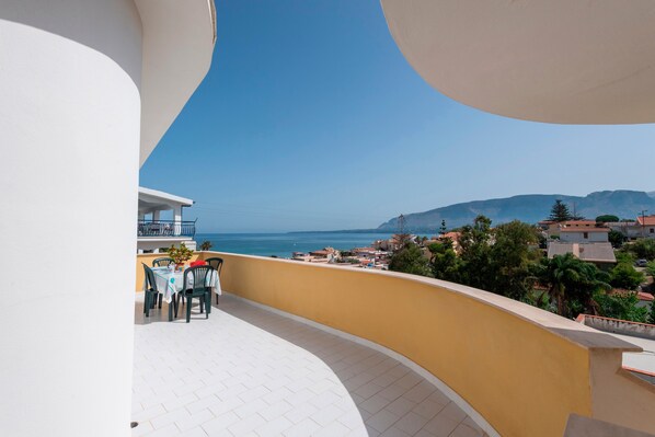 A balcony with a view of the sea and mountains, a tiled floor, and a yellow railing.