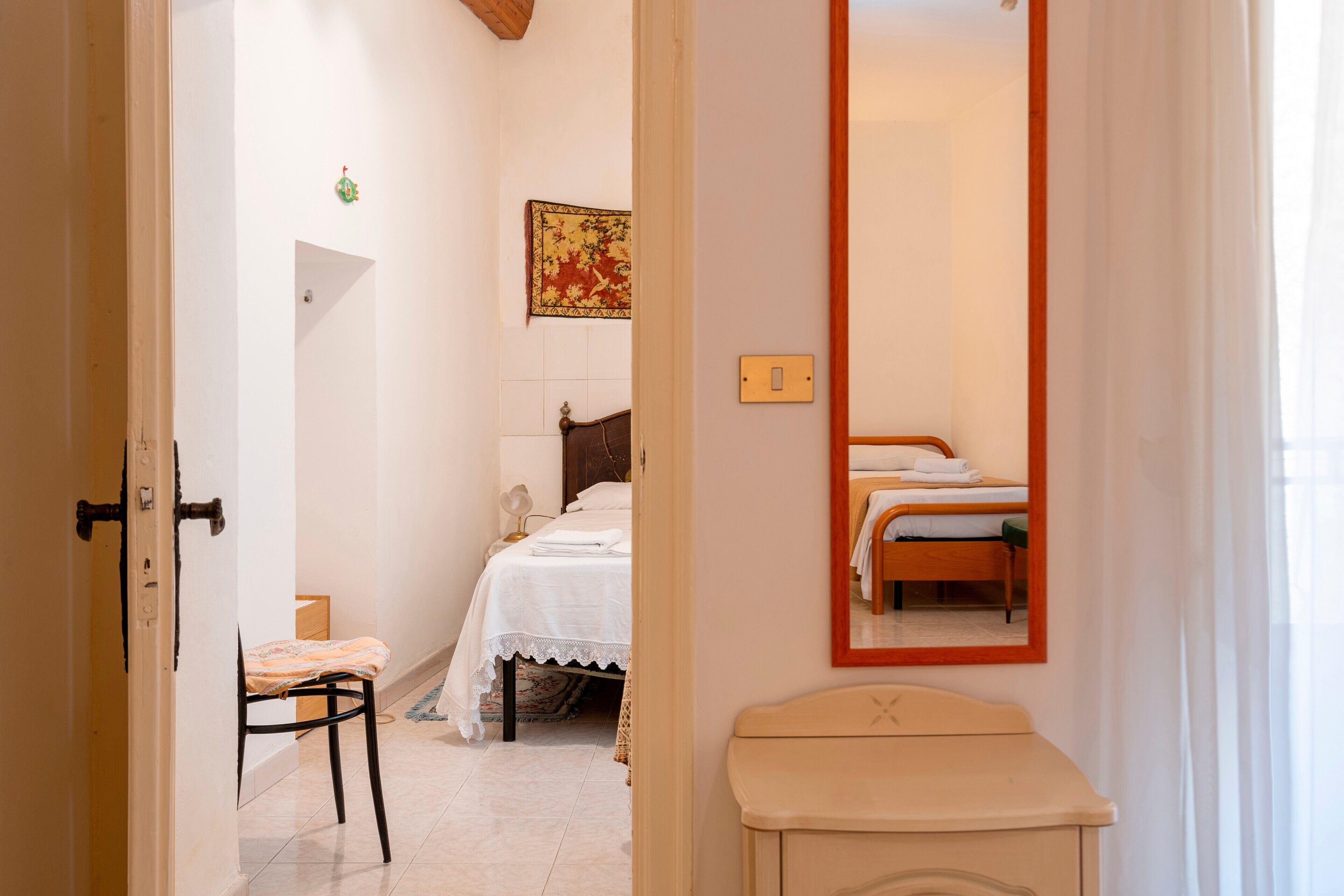 A bedroom with a bed, a chair, a bedside table, a mirror, and a wall-mounted light fixture.