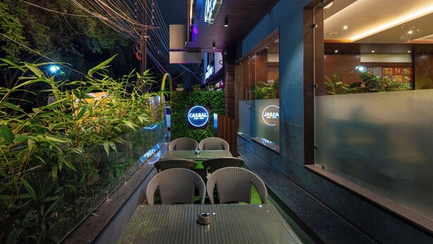 Un espace de restauration en plein air, avec des chaises et des tables en osier, entouré de verdure et délimité par une paroi en verre.