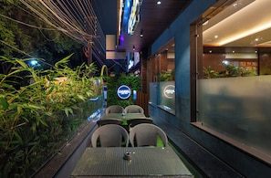 An outdoor dining area with wicker chairs and tables, surrounded by greenery and a glass wall. - Hotel Aston (Indore)