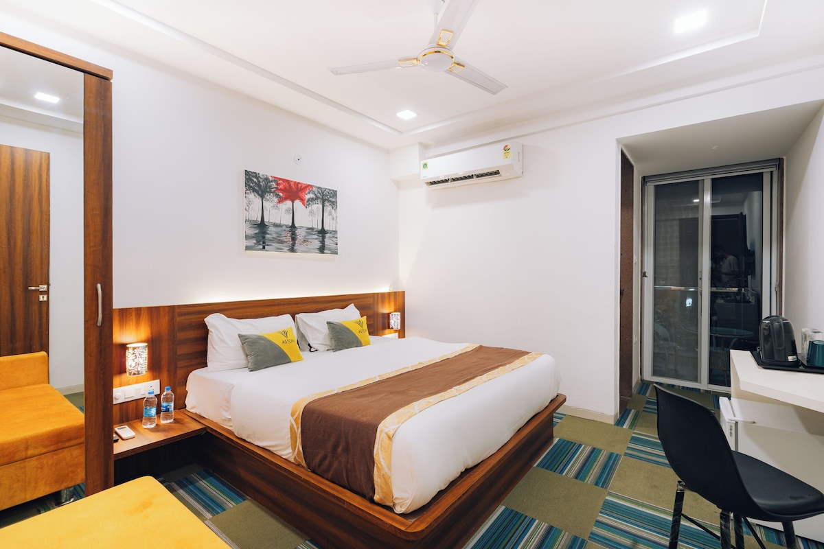 A hotel room with a bed, bedside table, chair, desk, and a view of the outside.