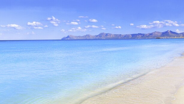 Una playa con agua azul cristalina y una cadena montañosa al fondo.
