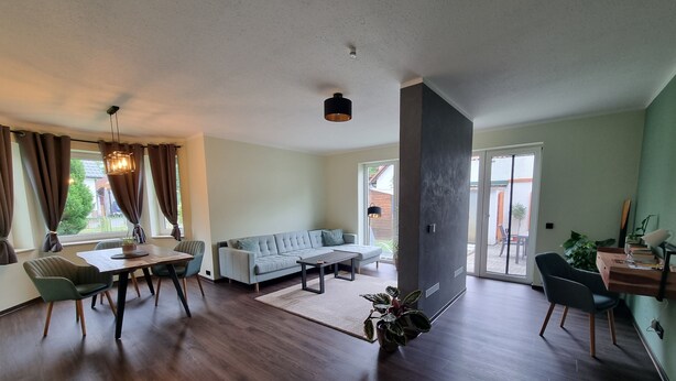 Een moderne woonkamer met een bank, eettafel en stoelen. Er is een groot raam met gordijnen, een plafondlamp en een plant.