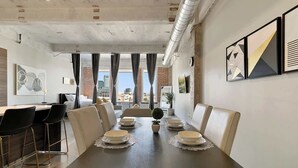 A dining area with a long table set for six, surrounded by chairs, with a city view through large windows.