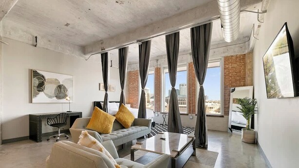 A modern living room with a sofa, armchairs, a coffee table, and a desk. There are large windows with curtains, a potted plant, and a painting on the wall.