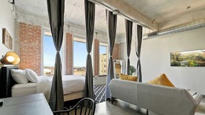 A hotel room with a bed, a sofa, a table, and a chair. There are curtains, a lamp, and a painting on the wall.