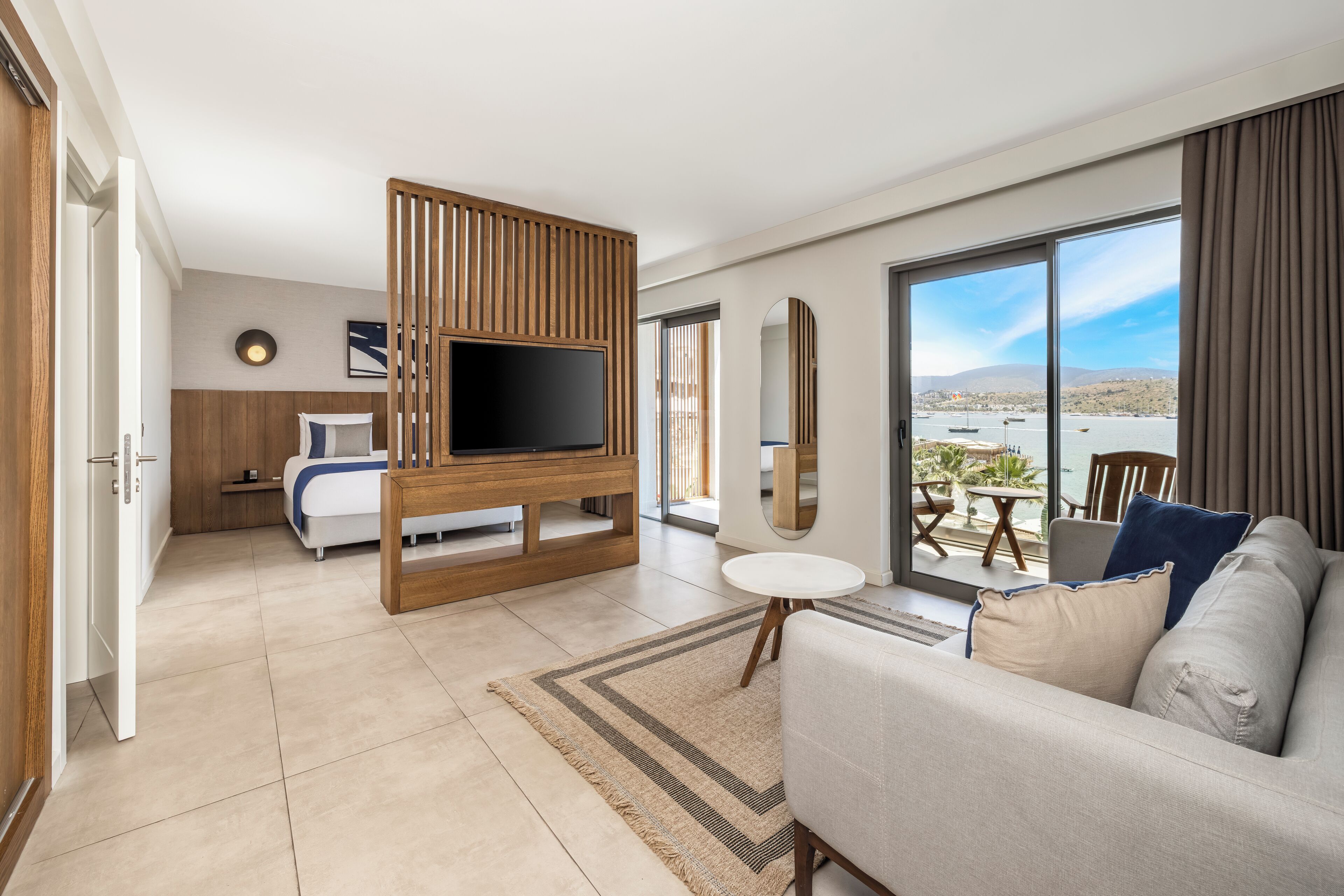 Habitación de hotel moderna con televisor grande, sofá, zona de comedor y balcón con vista al agua.