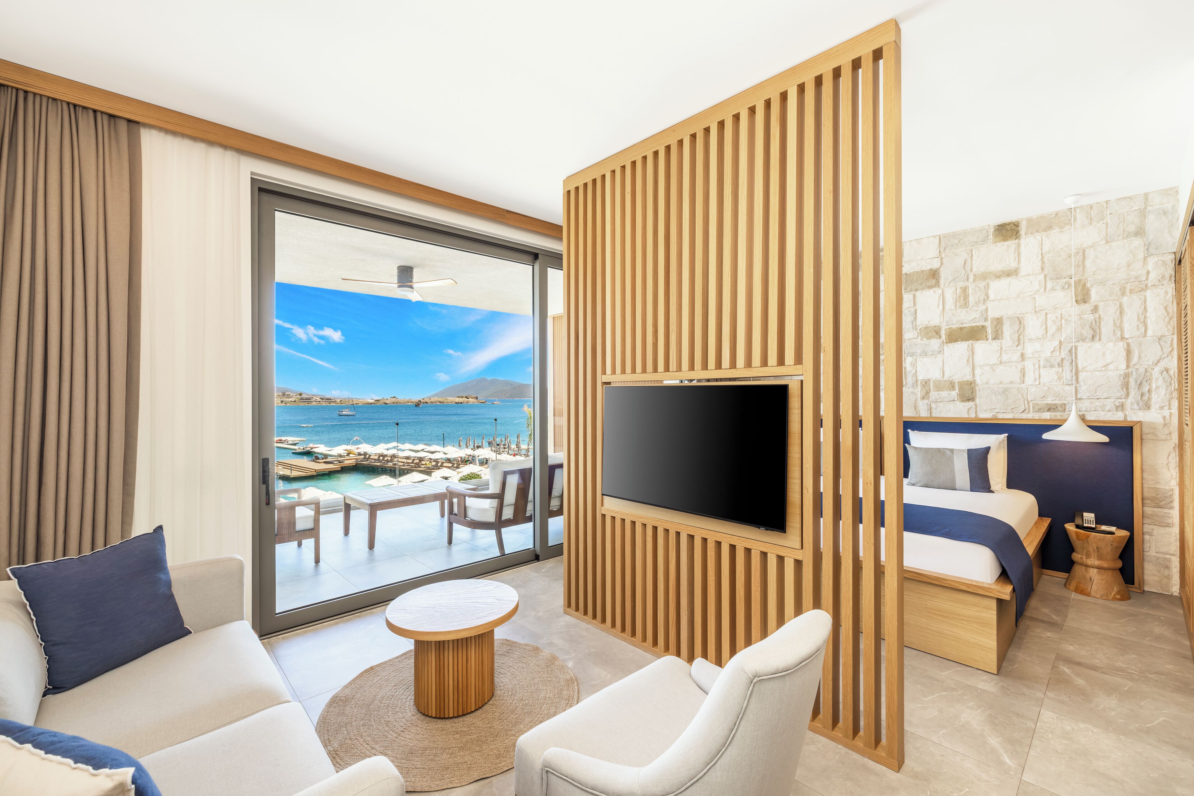 A modern hotel room with a large TV, comfortable seating, a view of the sea, and a stone wall.