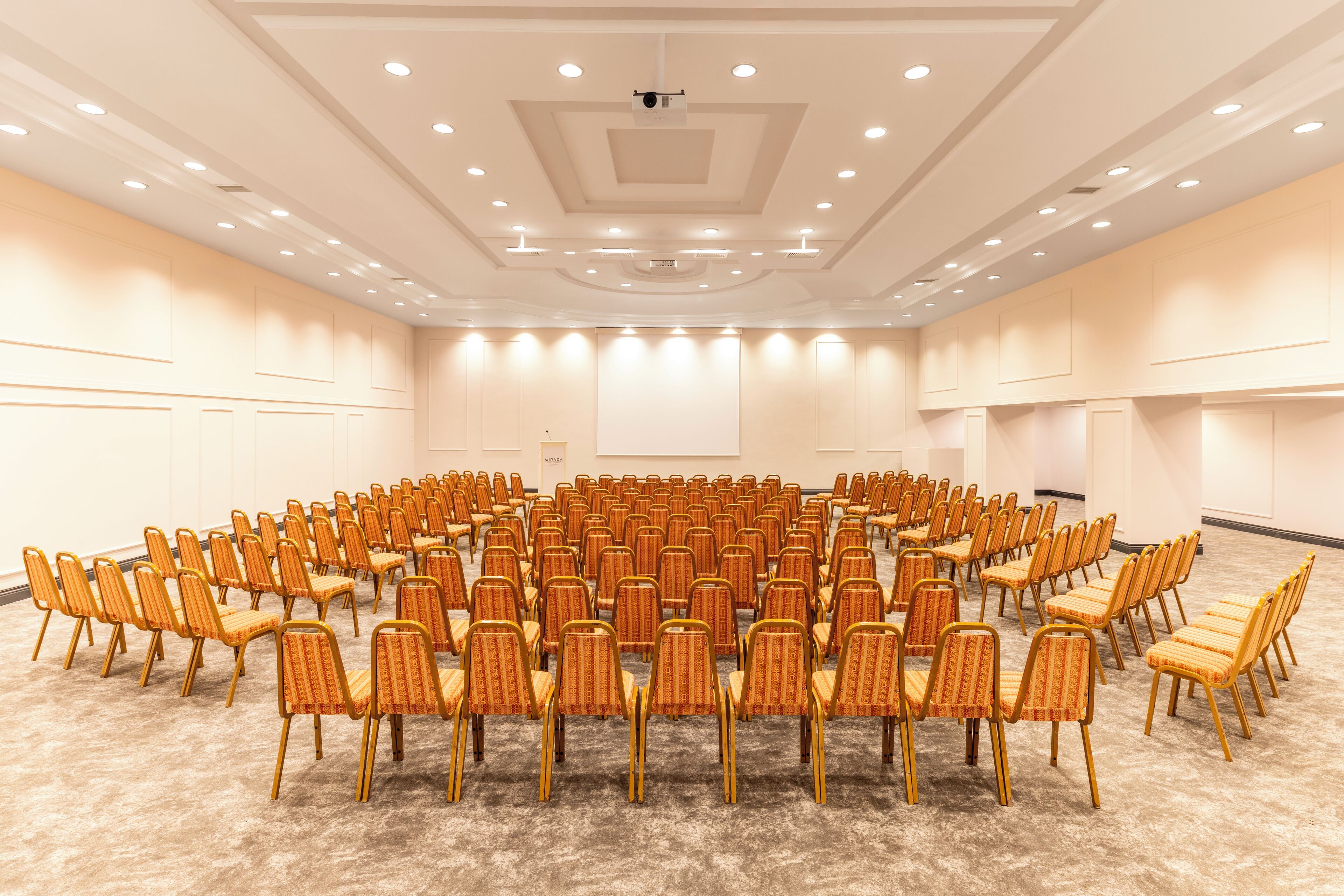 Un amplio salón de conferencias con filas de sillas y una pantalla de proyector.
