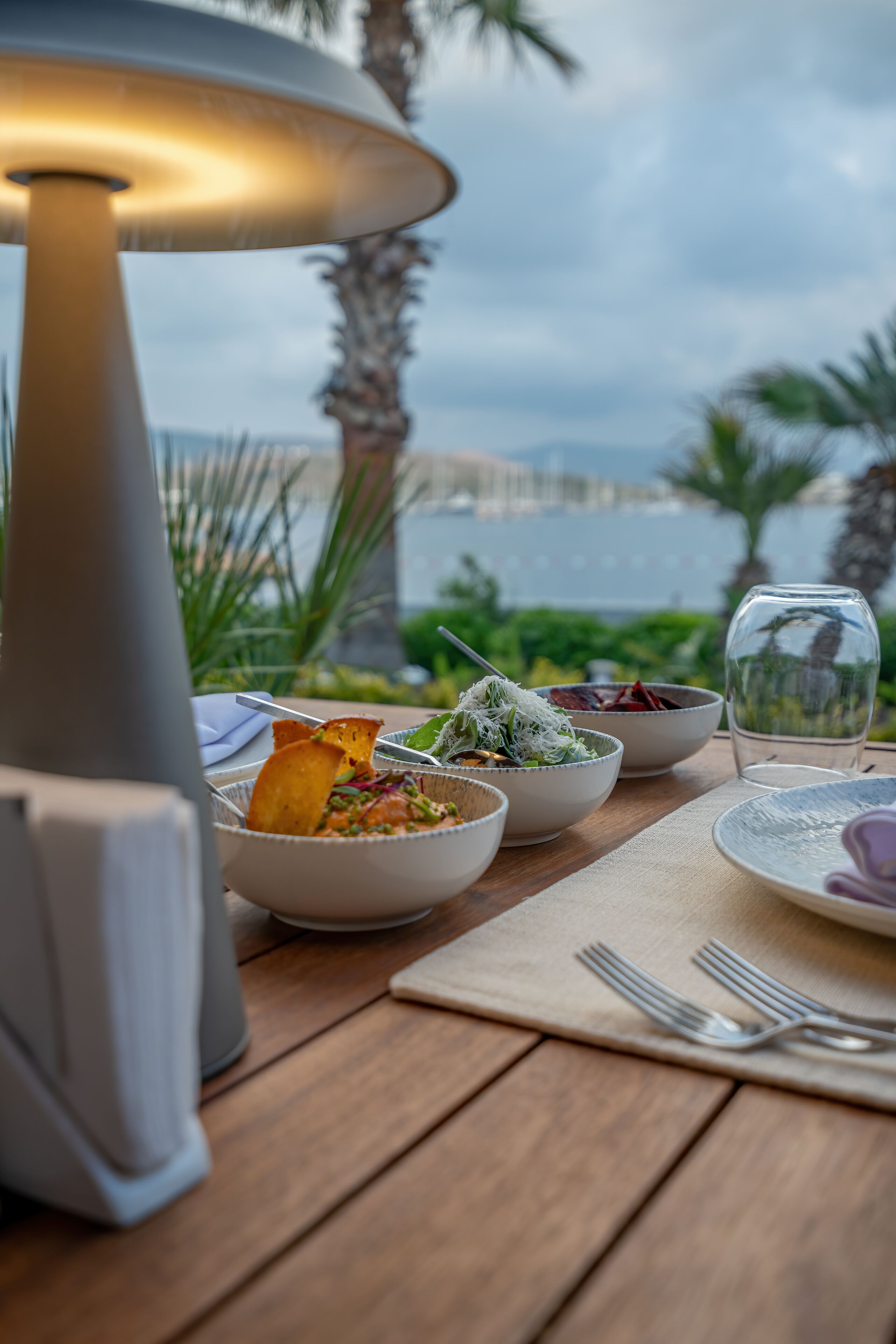 Una mesa dispuesta con varios platos, una lámpara y un vaso de agua, con vista a un cuerpo de agua y palmeras.