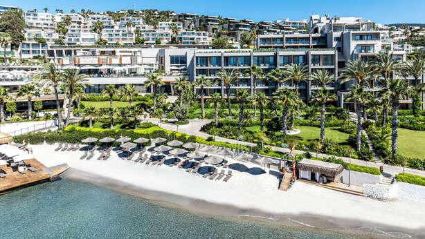 Een resort met een strand, palmbomen en moderne gebouwen.