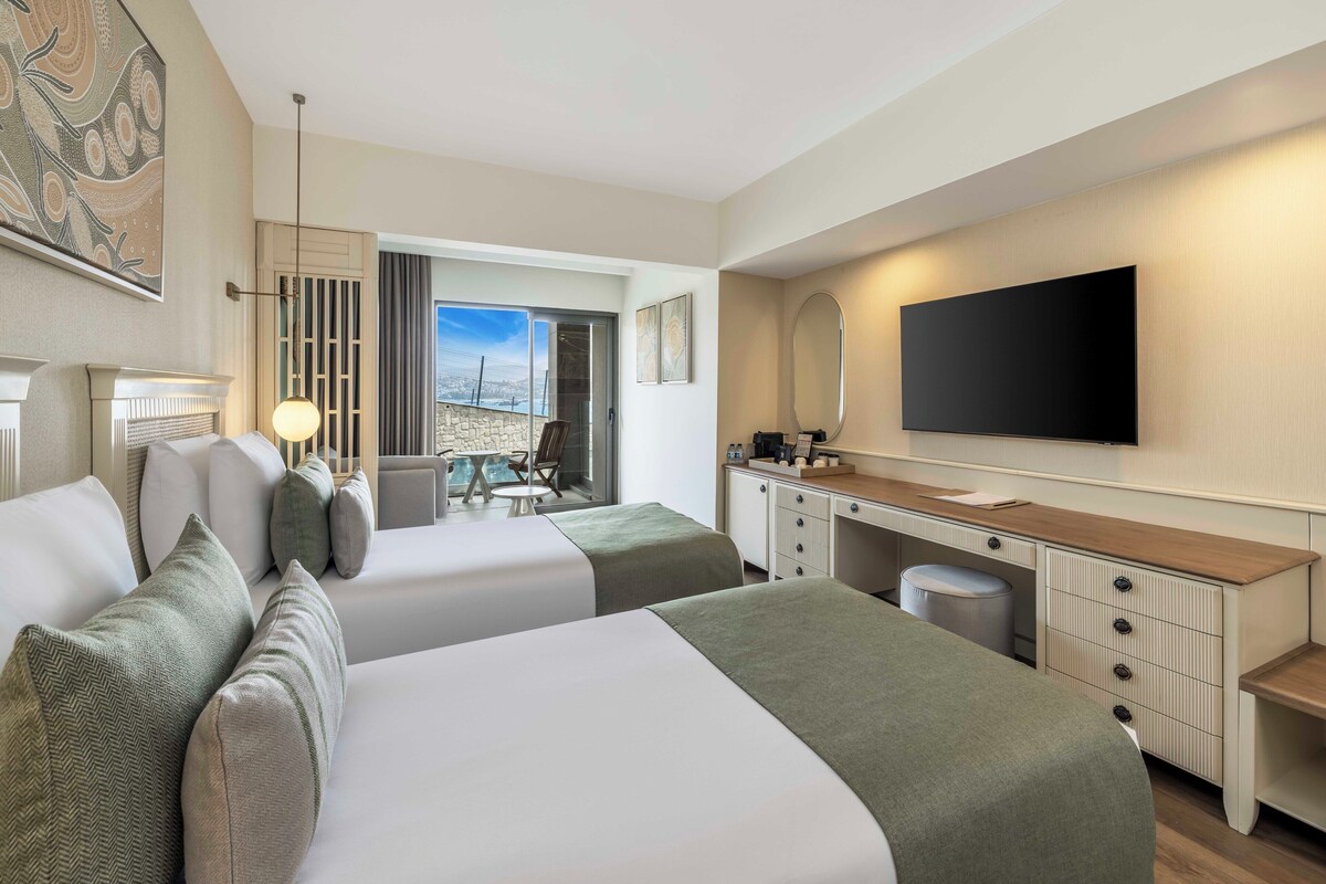 A hotel room with two beds, a TV, a desk, and a view of the city.