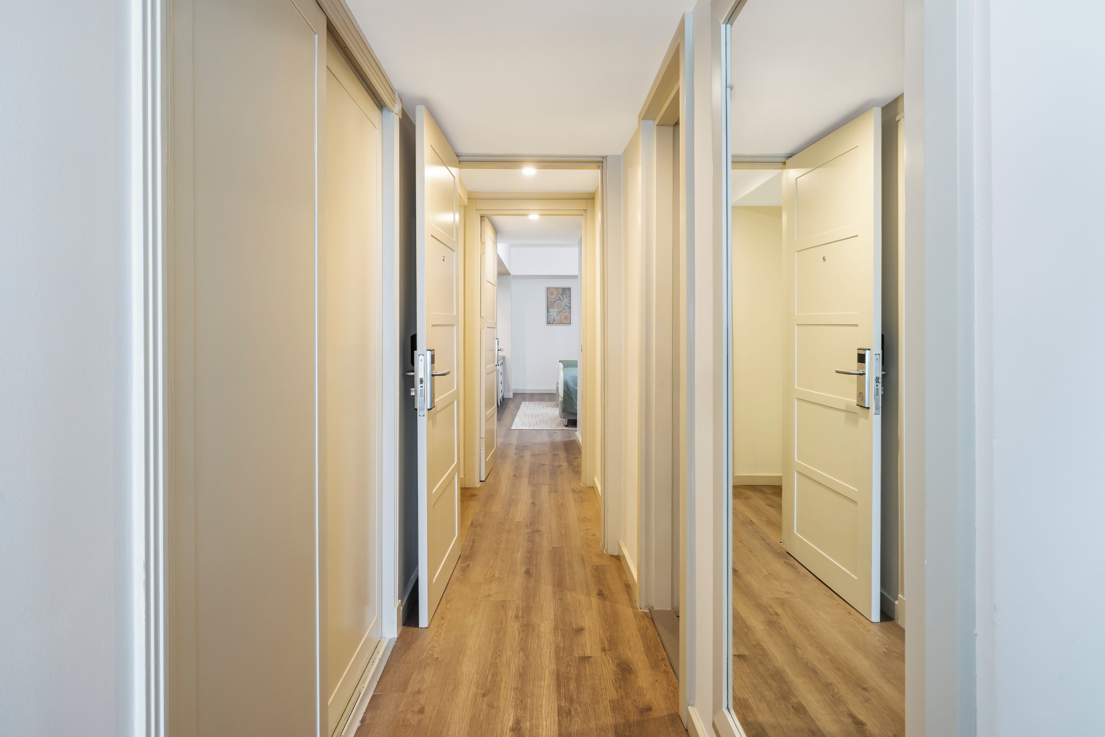 A narrow hallway with wooden flooring and white walls, leading to a room with a bed and a small table.