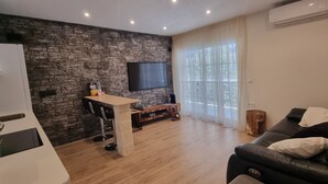 Ein modernes Wohnzimmer mit Steinwand, Flachbildfernseher, Holztisch, schwarzem Ledersofa und großem Fenster mit transparenten Vorhängen.