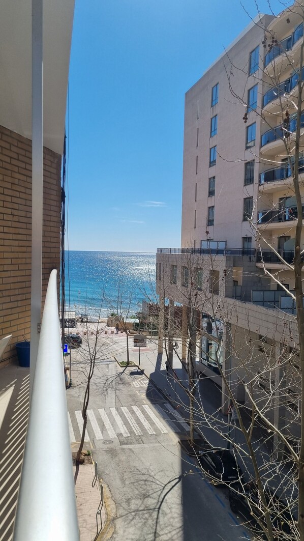A balcony overlooking a beach with a view of the sea and a building with multiple balconies.