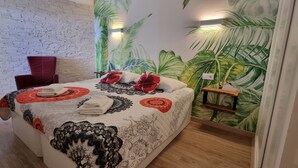 Ein Hotelzimmer mit Backsteinwand, einem großen Bett mit gemusterten Bettbezügen, einem roten Sessel und einem Wandbild mit tropischen Blättern.