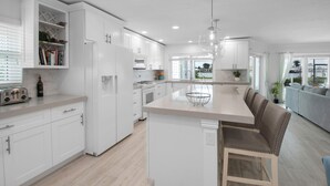 A modern kitchen with white cabinetry, a central island with seating, and a view of the outdoors through large windows. - CAPRI PARADISE!  Beautiful waterfront home.  Private heated pool and dock (Naples)
