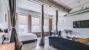 A modern hotel room with a large bed, a blue sofa, and a city view through large windows. - Downtown Loft: Walk to Convention (Dallas)