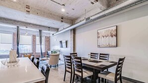 A modern dining area with a long table, wooden chairs, and a white countertop. - Downtown Loft: Walk to Convention (Dallas)