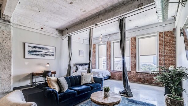 A modern living room with a dark blue sofa, a round coffee table, and a large window with a view of the city. - Downtown Loft: Walk to Convention (Dallas)