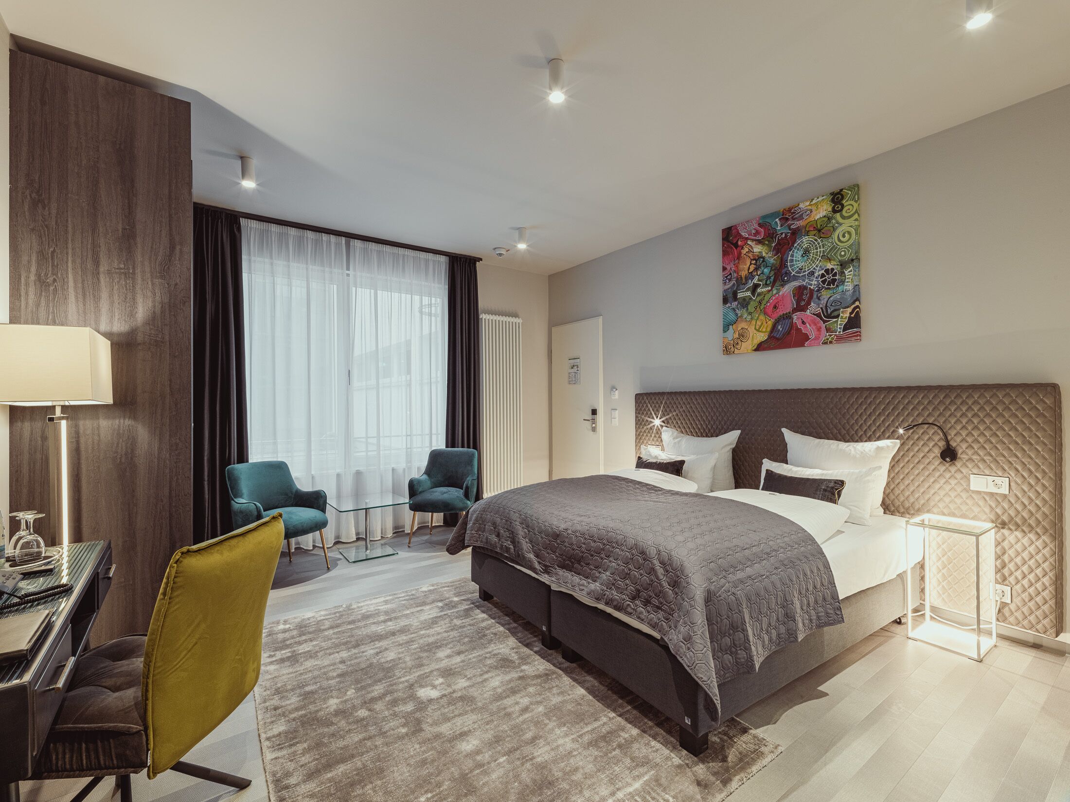 Una habitación de hotel moderna con una cama grande, un escritorio con una silla, una mesita con dos sillas y una obra de arte colorida en la pared.
