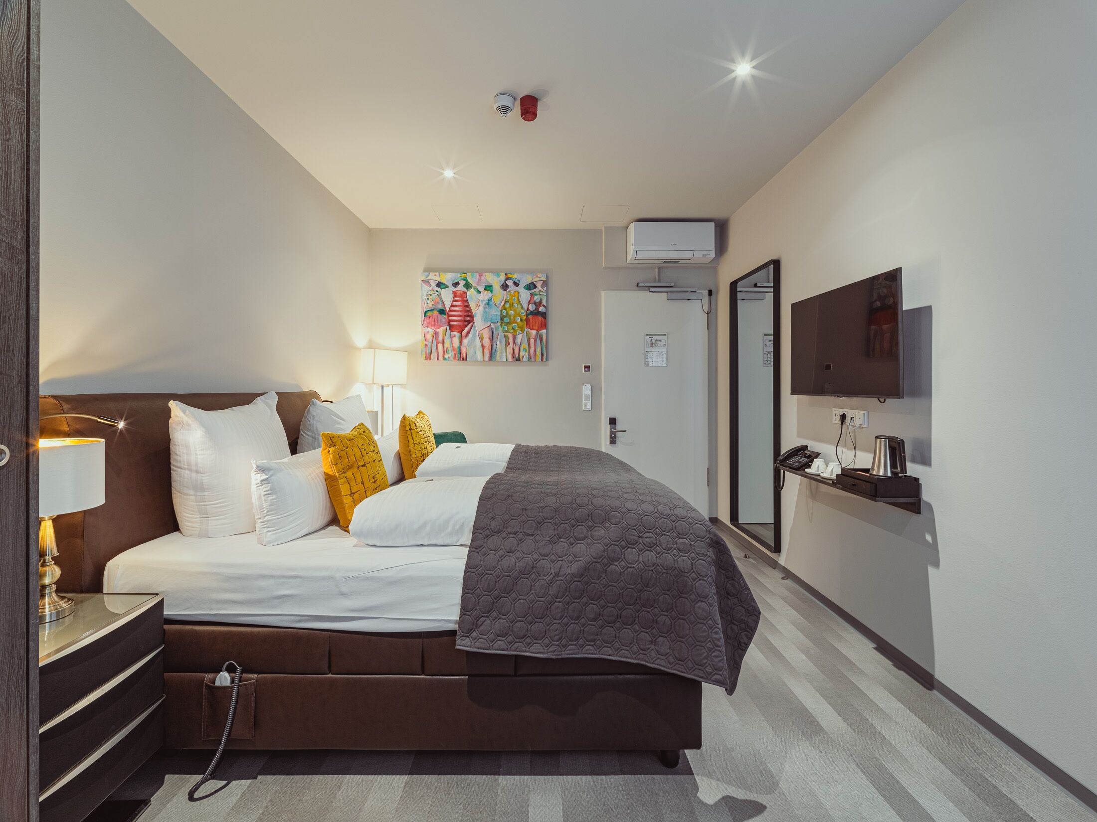 A modern hotel room with a large bed, bedside table, lamp, and a flat-screen TV mounted on the wall.