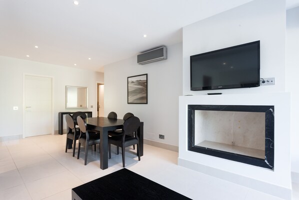A modern living room with a dining area, a flat-screen TV mounted above a fireplace, and a framed picture on the wall.