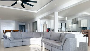 A modern living room with a grey sectional sofa, a ceiling fan, and a kitchen area in the background. - Coastal luxury paradise (Rosarito)