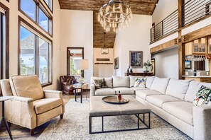 A spacious living room with a high ceiling, large windows, a chandelier, and a cozy seating area.