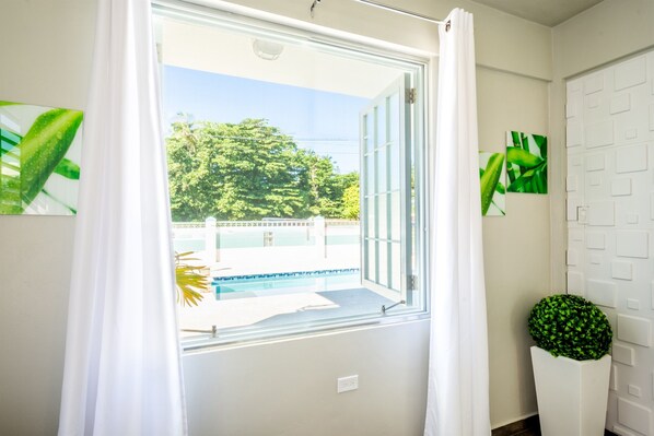 A room with a large window, a potted plant, and a white door with a geometric pattern. - 2 units | Sleeps 12 | Beach | Pool | Sunsets | Fun (Aguada)