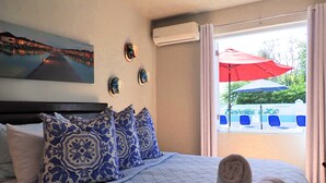A neatly made bed with blue and white patterned pillows, a rolled towel, and a painting of a coastal scene on the wall.