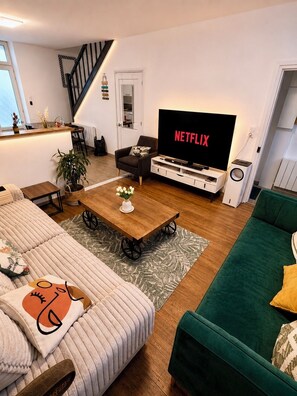 En moderne stue med sofa, grøn sofa, sofabord, et fjernsyn der viser 'NETFLIX' og en trappe.