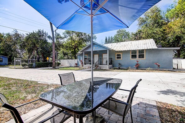 An outdoor patio with a glass table and chairs, a blue umbrella, and a house in the background.