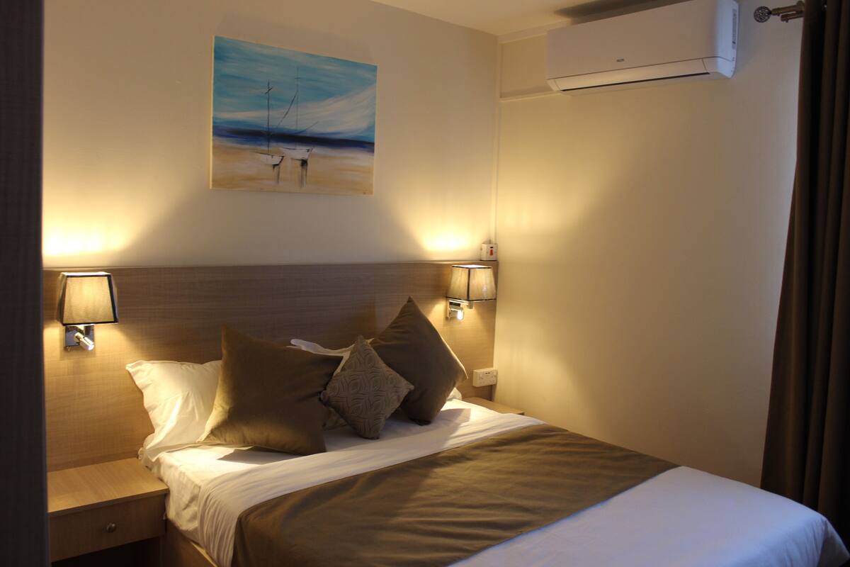 A hotel room with a neatly made bed, two bedside lamps, a painting on the wall, and an air conditioning unit.