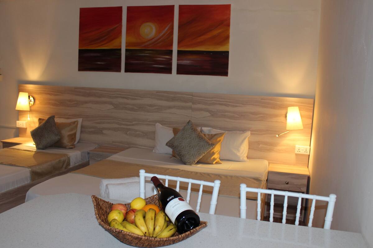 A hotel room with two beds, a dining table with fruit and wine, and artwork on the wall.