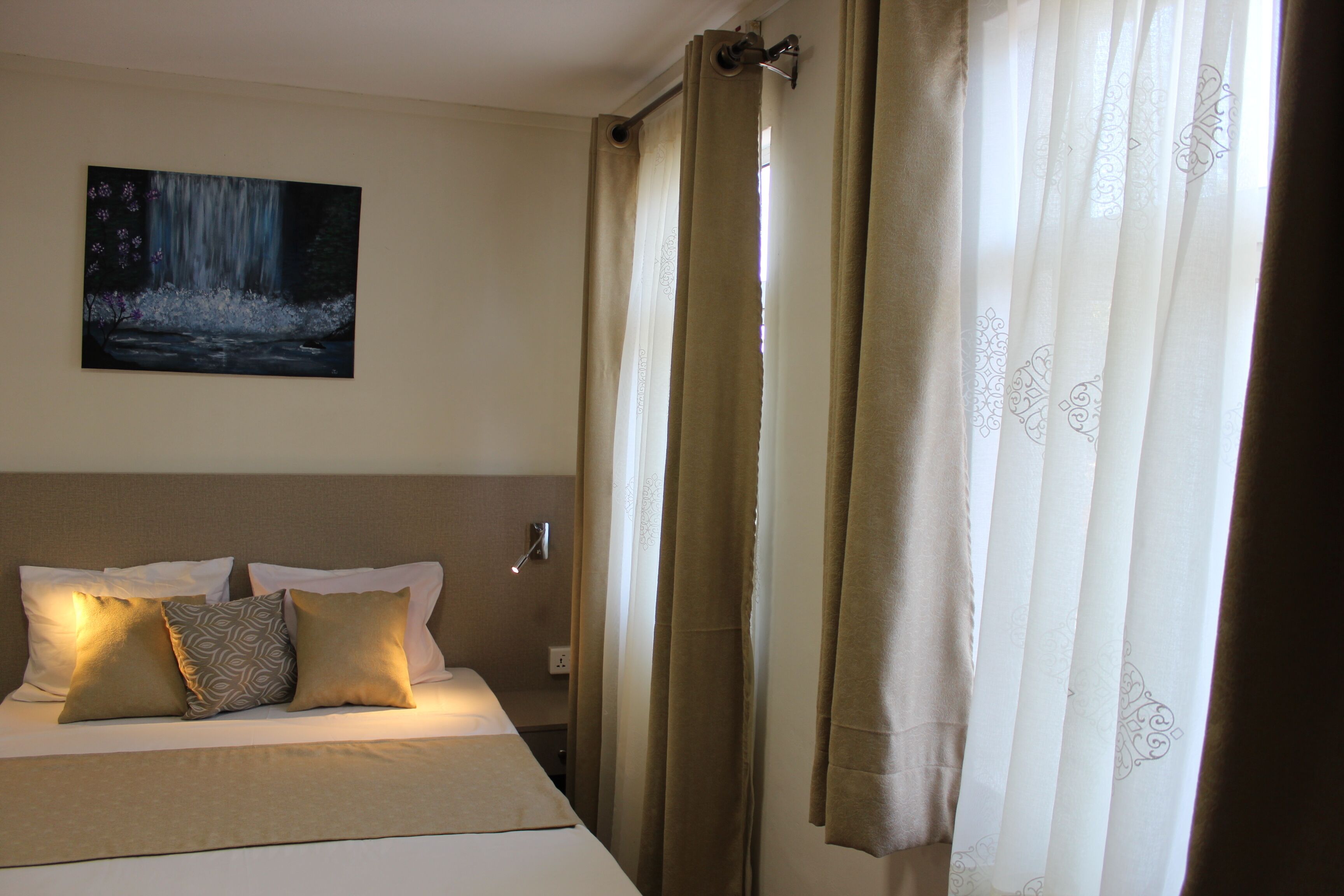 Una habitación de hotel con cama, almohadas, un cuadro en la pared y cortinas.