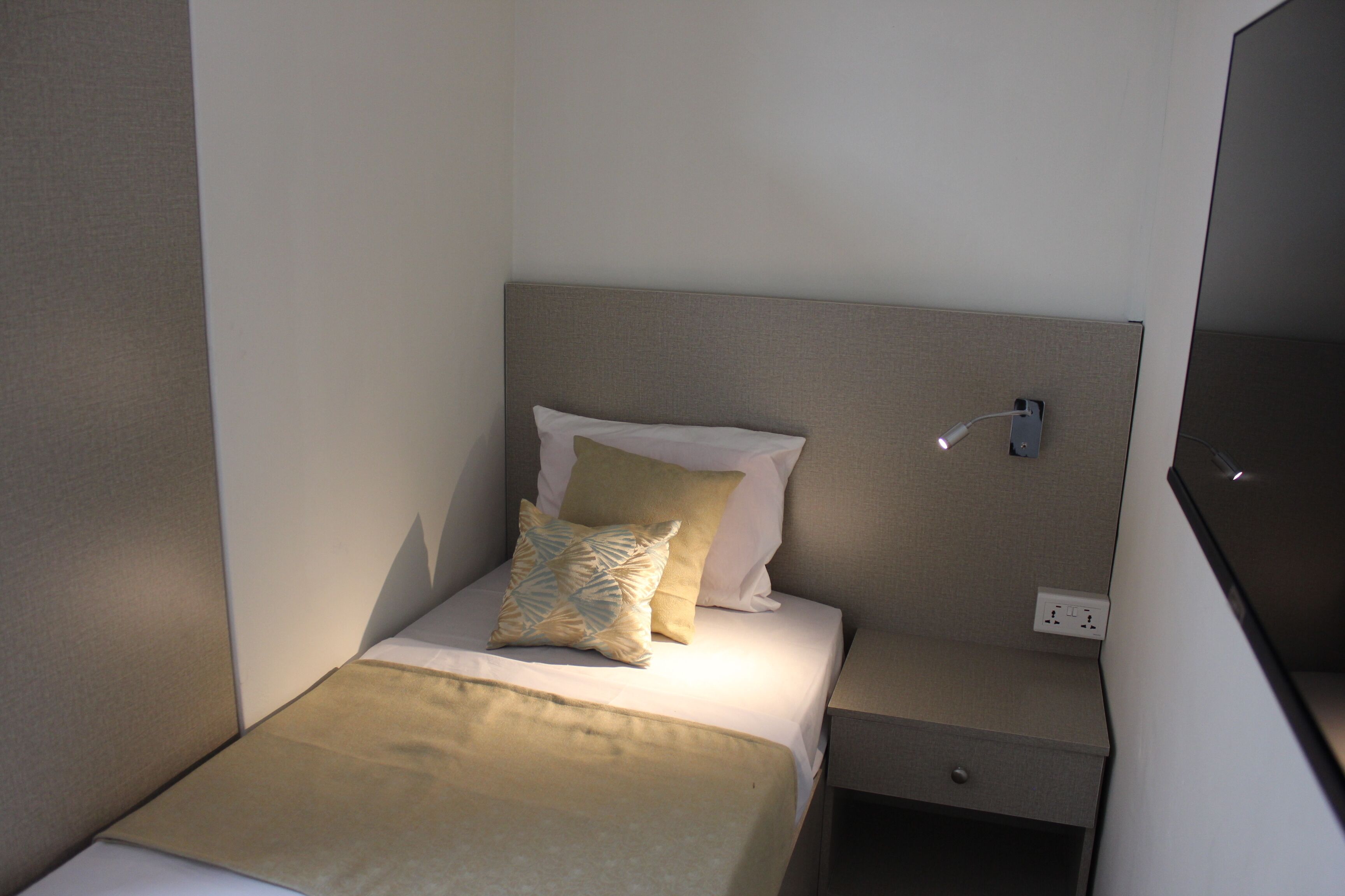 A neatly made bed with a beige bedspread and pillows, a bedside table with a lamp, and a power outlet on the wall.