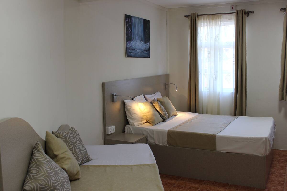 A hotel room with two beds, a nightstand, a wall-mounted lamp, a painting of a waterfall, and a window with curtains.