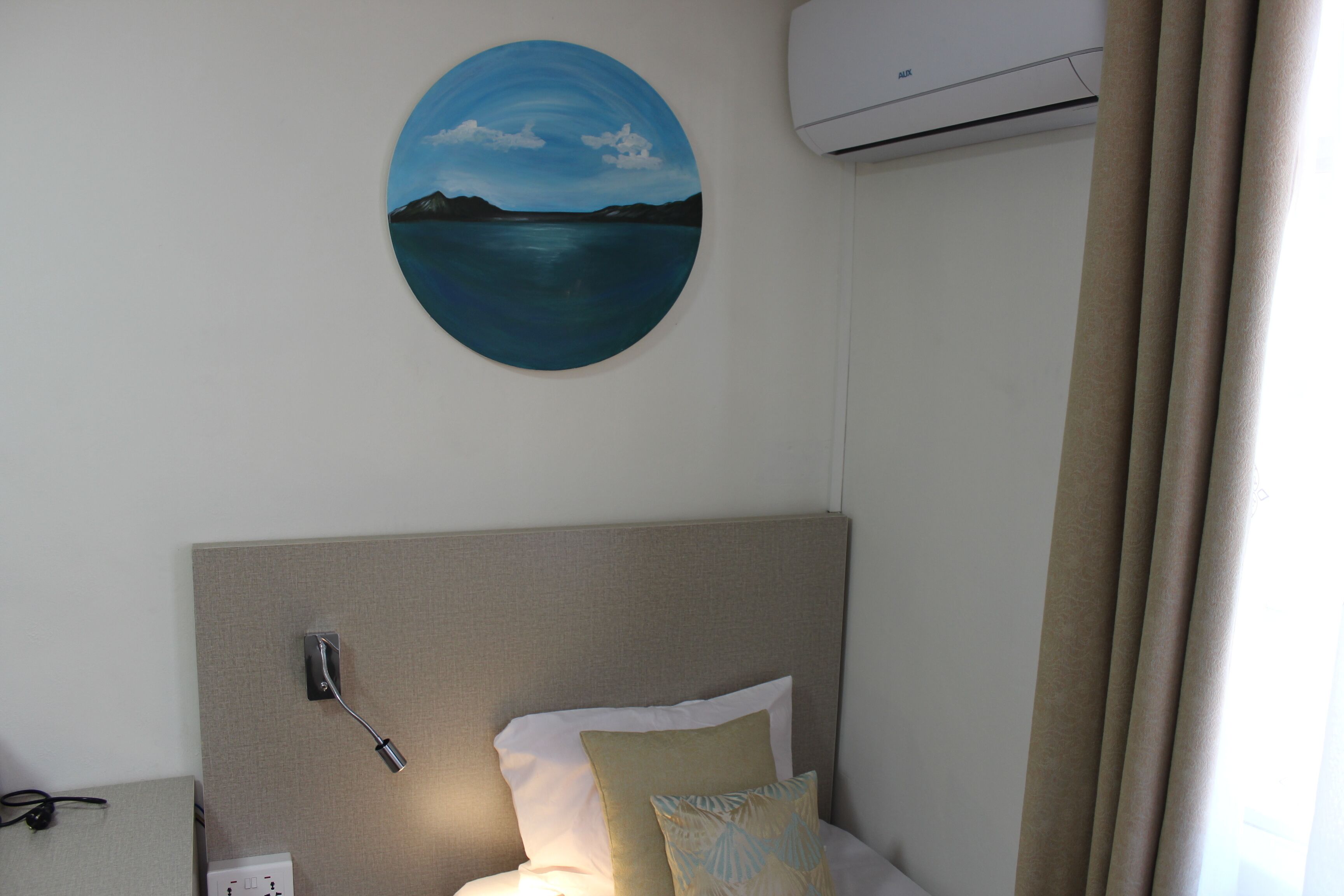 Una habitación de hotel con cama, aire acondicionado de pared, un cuadro de un paisaje y un enchufe.