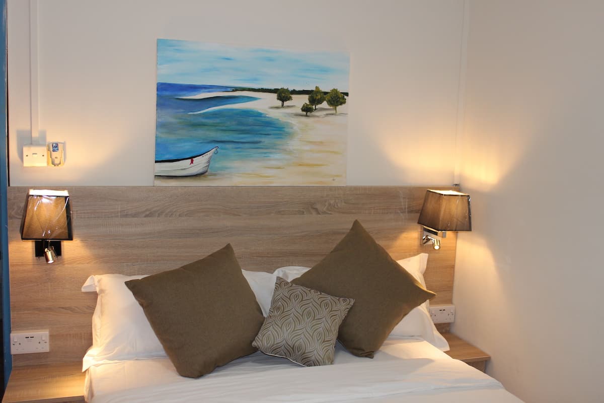 A neatly made bed with two pillows, a painting of a beach scene above it, and two wall-mounted lamps on either side.