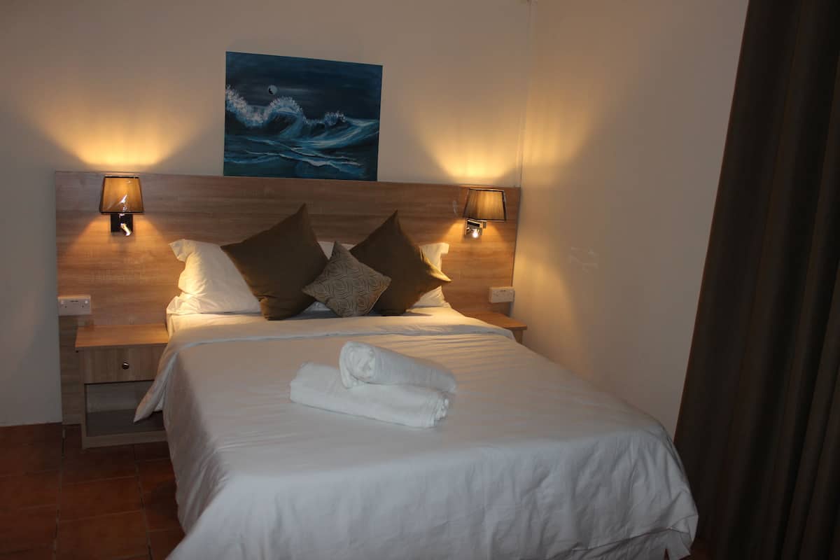 A neatly made bed with white linens and a rolled towel, two wall-mounted lamps, and a painting of a wave on the wall.