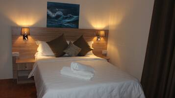 A neatly made bed with white linens and a rolled towel, two wall-mounted lamps, and a painting of a wave on the wall.