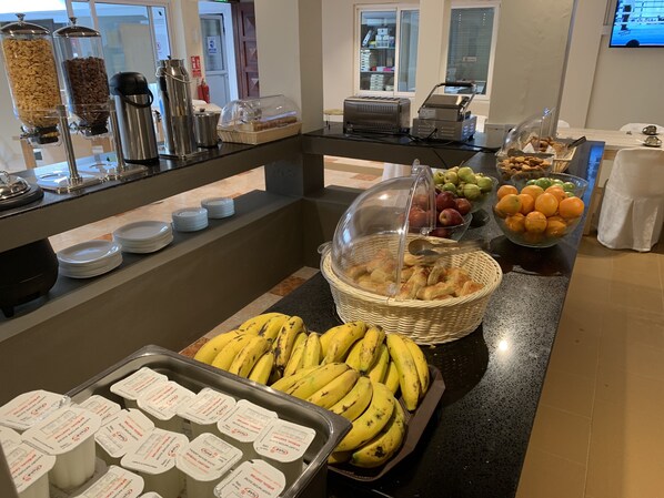 Zona de bufé con diversas opciones de comida, como plátanos, manzanas, naranjas y pasteles.