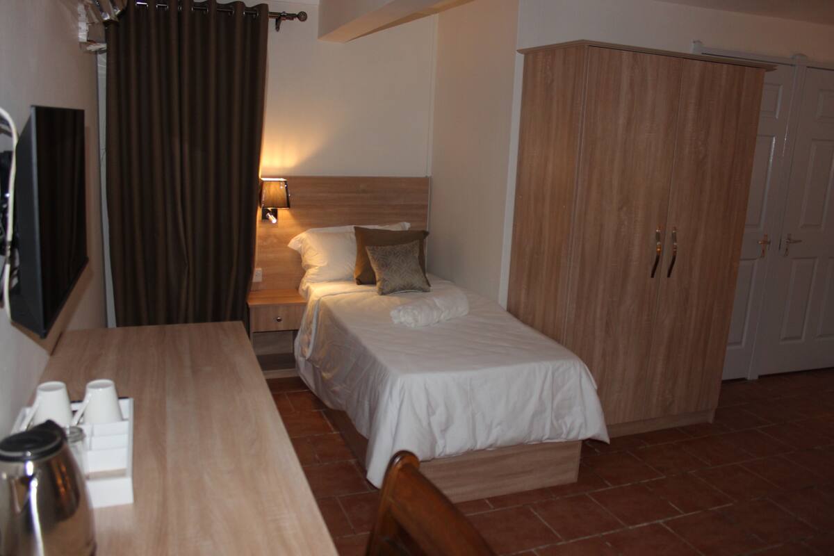 A bedroom with a single bed, a wooden wardrobe, a bedside table with a lamp, and a table with cups and a thermos.