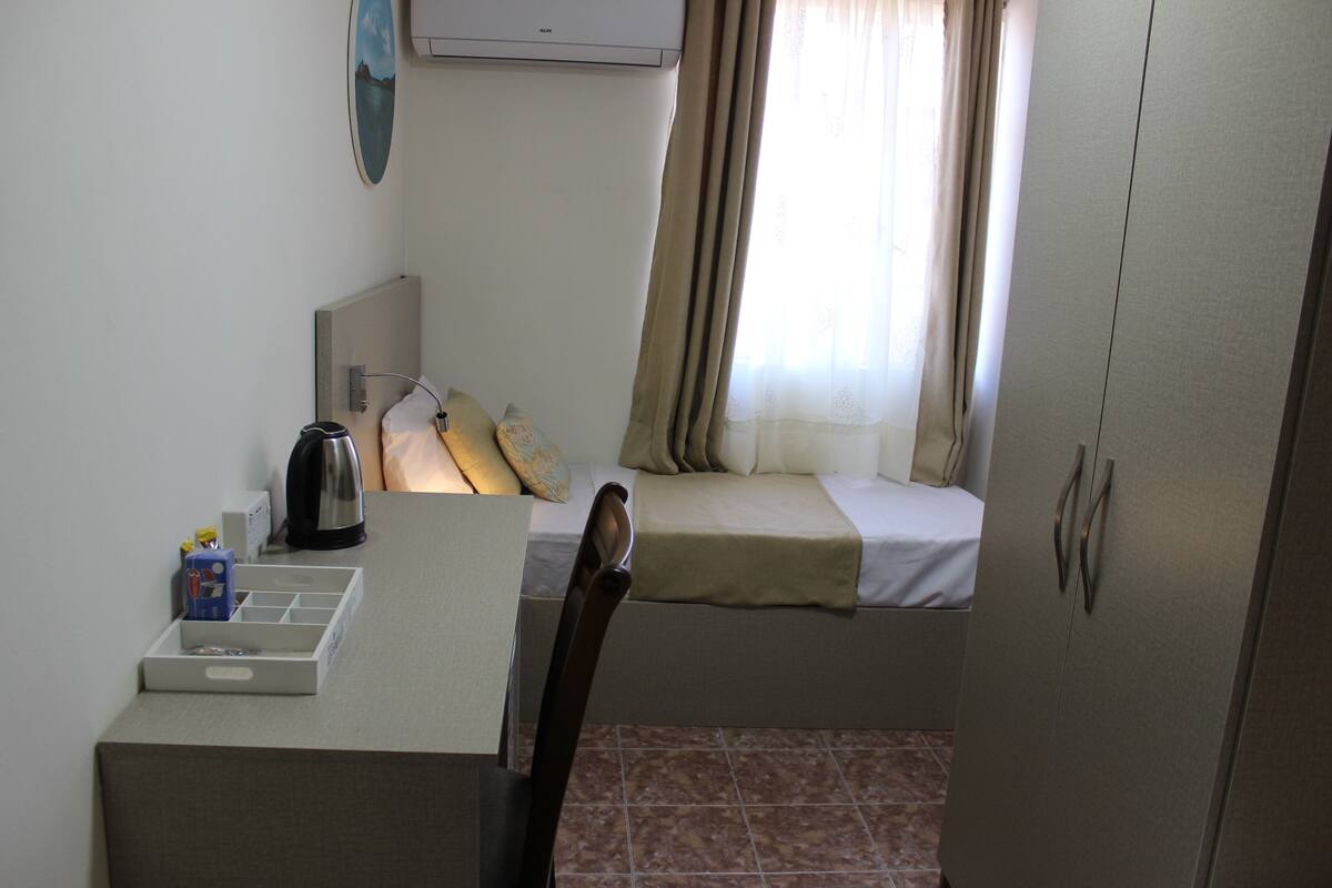 A small room with a bed, a desk with a kettle, a chair, and a wardrobe.