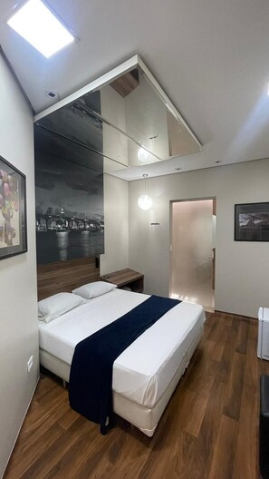 A modern bedroom with a bed, a nightstand, a mirror, and a view of a cityscape through the window. - Living Hotel Flex Inn (São Paulo)