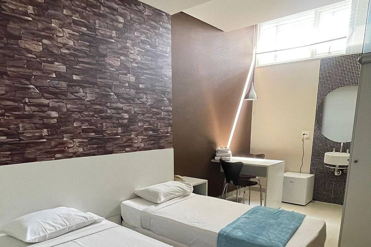 A hotel room with two beds, a desk, a chair, a small table, a mirror, and a bathroom with a sink and mirror.