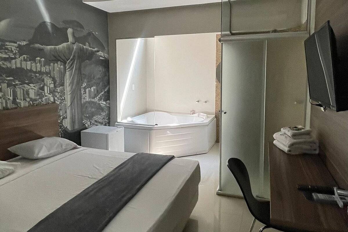 A modern hotel room with a large bed, a desk, a chair, a vanity with a mirror, and a small seating area with a table and chairs.