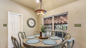 A dining area with a round table, six chairs, a decorative hanging light, a clock, and a window with a view of the outdoors.
