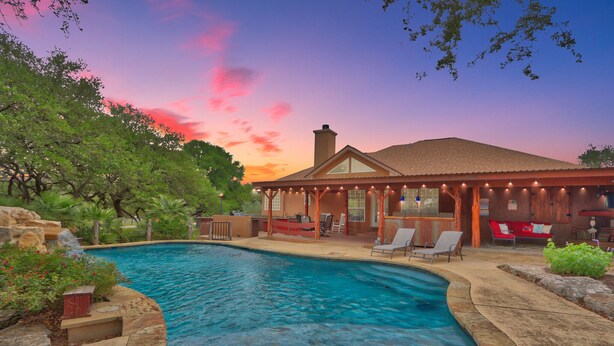 A pool area with a house in the background, surrounded by trees and a sunset sky.